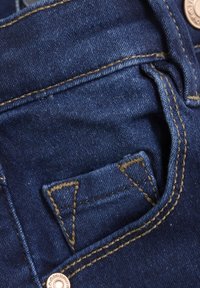 Tamno plavi denimski materijal s vidljivom džepom, naglašen žutim šavom i metalnim dugmetom. Ima trokutasti oblik i teksturu džepa.