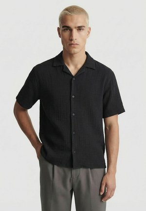 Giovane uomo con capelli biondi corti, indossa una camicia nera a maniche corte con bottoni e pantaloni grigi su uno sfondo chiaro e uniforme.