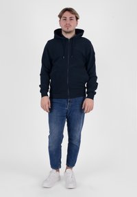 Felpa con zip di colore blu navy con cappuccio regolabile e polsini a costine, abbinata a jeans a sigaretta blu chiaro e sneakers bianche. Sfondo semplice.