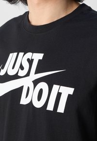 Osoba ubrana w czarną koszulę z dużym białym napisem "JUST DO IT" oraz białym logo Nike na klatce piersiowej.