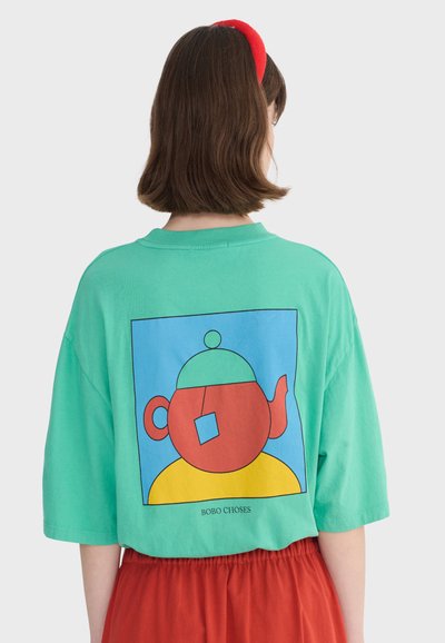 Femme aux cheveux bruns portant un bandeau rouge, un t-shirt vert avec un motif abstrait de théière et un pantalon rouge, debout devant un fond uni.