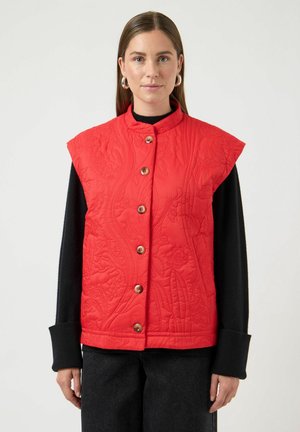 Femme portant un gilet matelassé rouge sans manches avec de gros boutons, par-dessus un haut noir à manches longues et un pantalon sombre, debout devant un fond uni.