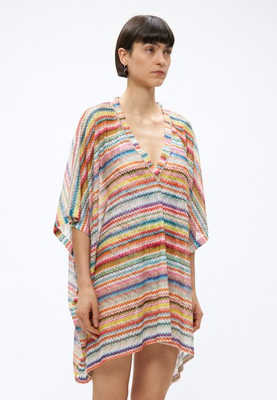 Γυναίκα με κοντά σκούρα μαλλιά που φοράει αέρινο φόρεμα με πολύχρωμο μοτίβο σε σχήμα ζιγκ ζαγκ, V-λαιμόκοψη και μανίκια μέχρι τον αγκώνα, στέκεται μπροστά σε απλό φόντο.