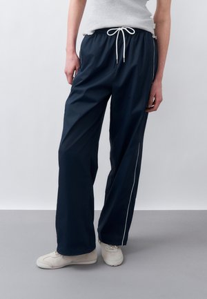 Άτομο που φοράει ναυτικό μπλε παντελόνια με φαρδιά πόδια, με λευκό κορδόνι και ρίγα, σε συνδυασμό με λευκά αθλητικά παπούτσια, στέκεται μπροστά σε απλό φόντο.