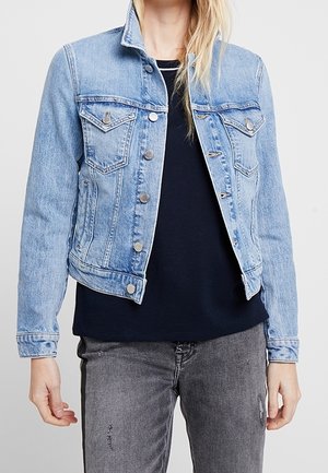 Persoon draagt een lichtblauwe spijkerjas over een zwart shirt en grijze spijkerbroek, staat met de armen ontspannen langs het lichaam.