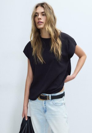 Mlada ženska z dolgimi valovitimi lasmi nosi ohlapno črno majico brez rokavov, svetlo modre kavbojke s črnim pasom in drži črno usnjeno jakno.