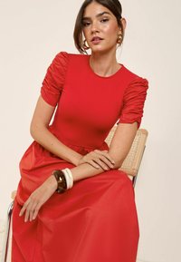 Robe rouge avec des manches courtes froncées, un corsage texturé et une jupe fluide. Le modèle porte un bracelet en cuir marron épais et deux bracelets blancs.