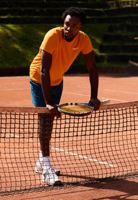 Tennis spiller i en orange Nike skjorte og blå shorts hviler mod et net, mens han holder en ketsjer med gule strenge på en grusbane.