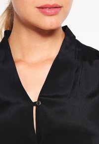Primo piano del viso inferiore e del collo di una persona che indossa una blusa nera di seta chiusa con un piccolo bottone.