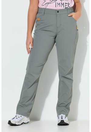 Vrouw draagt lichtgrijze rechte broek met zijzakken en witte sneakers, staand met één hand in de zak.