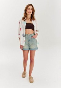 Biela vyšívaná košeľa s farebnými vzormi, hnedý crop top, svetlomodré riflové šortky s vyhrnutými manžetami a béžové slip-on topánky.