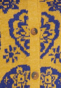 Stickad cardigan i gult med ett blått blommönster, med en central knäppning och textil med struktur.