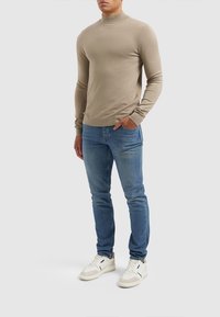 Beige, slim-fit, lange mouw trui met een opstaande kraag, gecombineerd met lichtblauwe jeans en witte sneakers met beige accenten.