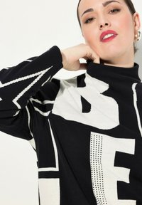 Maglione lavorato a maglia in bianco e nero con collo alto, caratterizzato da un testo grafico audace e motivi a zigzag a contrasto in morbido tessuto texturizzato.