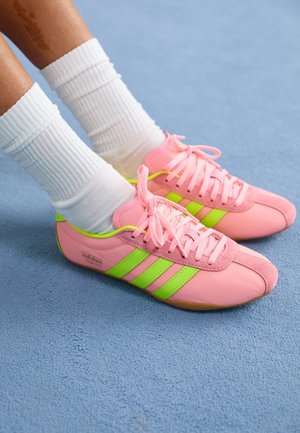 Fødder iført hvide crew-sokker og pink Adidas Tokyo sneakers med neongrønne striber på blåt tæppe.