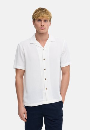 Junger Mann mit blondem Haar, der ein weißes kurzärmeliges Hemd und dunkle Hosen trägt, steht vor einem einfarbigen hellen Hintergrund.