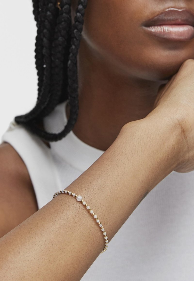 Bracciale in oro e perle su un polso, con delicate perle rotonde e una superficie liscia, che si abbina al tono della pelle. Design semplice.