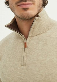 Pull beige zippé avec un col haut, en tissu texturé, et doublure intérieure marron. Présente une fermeture éclair en métal avec une tirette en cuir.