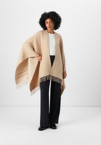 Beige poncho med frynser, laget av myk stoff, har åpen front og vinklet ermer, brukt over en hvit topp og mørke bukser.