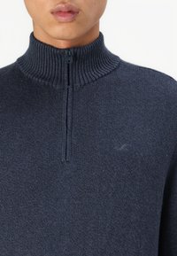 Marineblauer Pullover mit hohem Kragen und Reißverschluss vorne, geripptem Kragen und weicher Textur. Besitzt ein kleines gesticktes Logo auf der Brust.