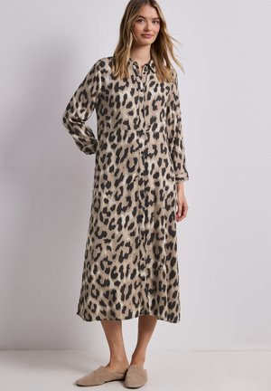 Frau steht und trägt ein beige-schwarzes Langarm-Midikleid mit Leopardenmuster und Knöpfen sowie beige Slip-On Schuhe.