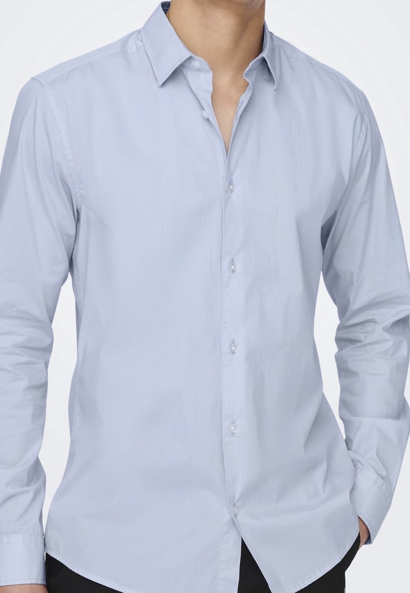 Camicia button-up a maniche lunghe di un blu chiaro con una texture morbida, colletto classico e orlo dritto. Presenta bottoni bianchi lungo il fronte.