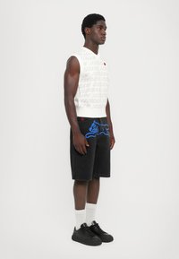 Hvid ærmeløs skjorte med gentaget tekstmønster, sorte denimshorts med blå kattegrafik, sorte sneakers og hvide sokker.