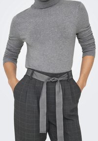 Pull gris à col roulé associé à un pantalon à carreaux foncé taille haute, avec une ceinture en tissu assorti nouée à l'avant.