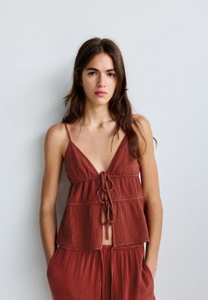 Mladá žena s dlhými hnedými vlasmi, ktorá nosí bezrukávový hrdzavočervený textúrovaný top s viazačkami vpredu a voľné nohavice v rovnakom odtieni na jednoduchom pozadí.