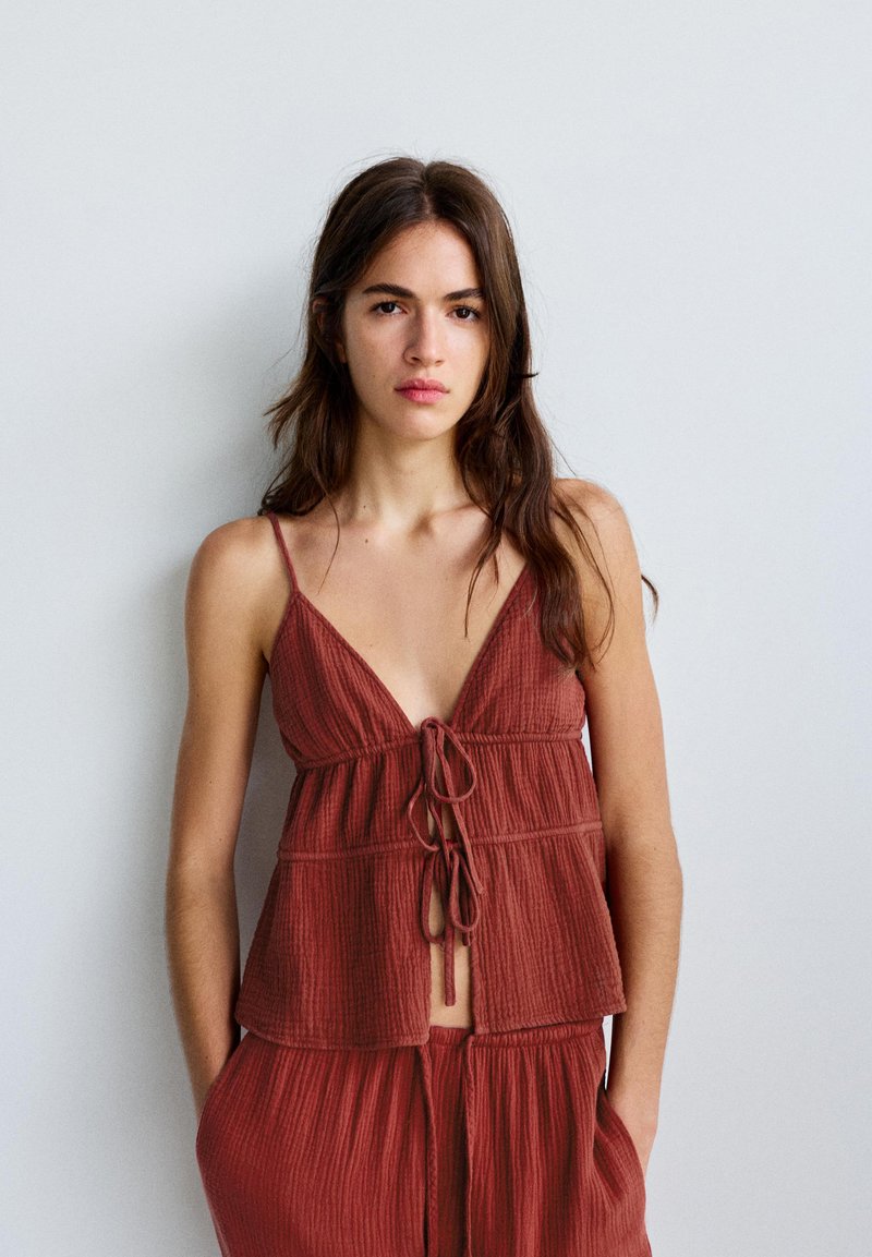 Jeune femme aux longs cheveux bruns portant un haut sans manches rouge rouille texturé avec des liens à l'avant et un pantalon ample assorti sur un fond uni.