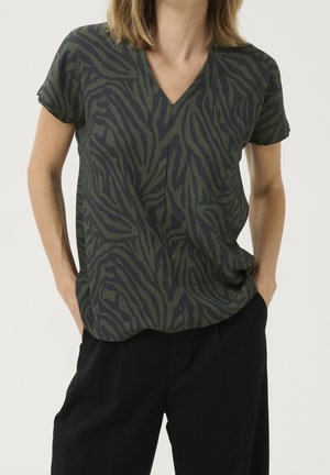 Personne portant un haut à manches courtes vert foncé à col en V avec des rayures zébrées abstraites noires et un pantalon noir, les mains dans les poches, sur un fond uni.