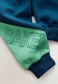 Zielona i granatowa bluza z ribbowymi mankietami; na zielonej części znajduje się duży, jasnoniebieski napis "BAKER". Widoczna miękka faktura tkaniny.