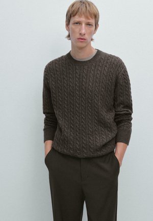 Maglione marrone a trama a treccia con scollatura rotonda, dotato di polsini e fondo a coste, abbinato a pantaloni scuri. Sfondo semplice.