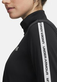 Black athletic top with a high collar and white "UNDER ARMOUR" logo tape along the shoulder. Smooth texture, fitted design.