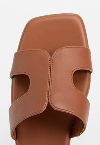 Sandale slide en cuir marron avec larges brides présentant des découpes symétriques et une couture cousue au centre.