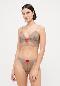 Spetsbralette och matchande tanga i ljusbrun, med scallopade kanter, tunna remmar och ett röd logoband. Texturerad spetsdetalj genom hela.