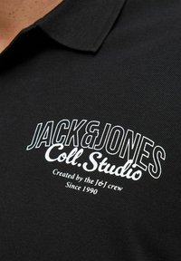 Schwarzes Poloshirt mit geripptem Kragen, strukturiertem Stoff und weißem, gesticktem Text: "JACK & JONES Coll. Studio" mit zusätzlichen Details darunter.