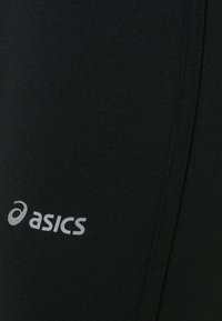 Leggings atletici neri con una texture liscia, caratterizzati da un logo Asics grigio sul lato inferiore sinistro.