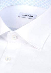 Col de chemise blanche et boutons supérieurs, étiquette de marque "seidensticker" visible à l'intérieur de l'encolure, posé sur un fond à motifs bleus et blancs.