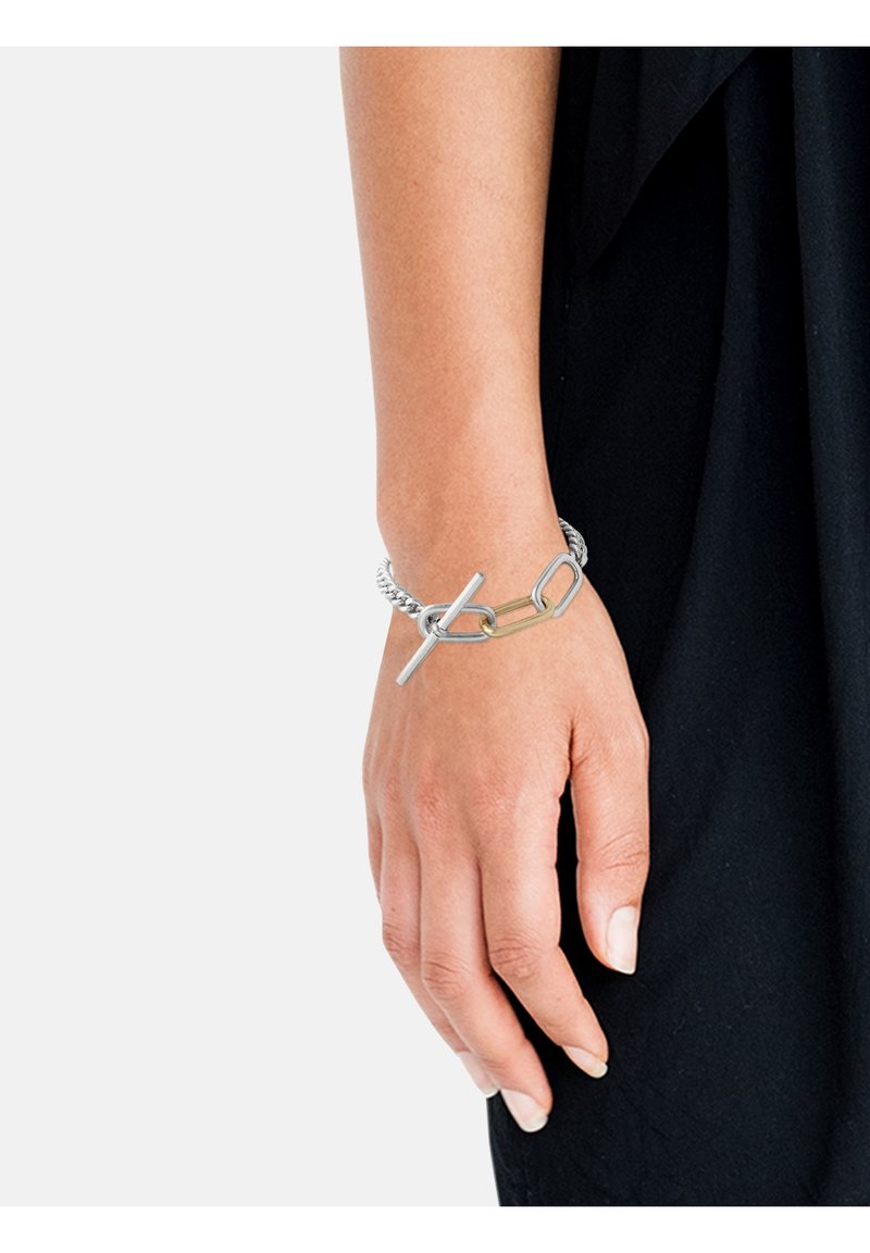 Main portant un bracelet chaîne en métal mêlé argent et or avec fermoir à bascule, reposant sur un fond en tissu noir.