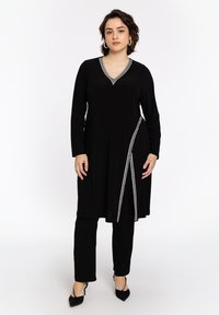 Zwarte lange tuniek met een V-hals en zilveren versieringen langs de nek en de zijkant. Gecombineerd met zwarte broeken en puntige hakken.