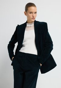 Žena ve černém sametovém blejzru a kalhotách s bílým žebrovaným topem s průhledným panelem, stojící na jednobarevném světlém pozadí.