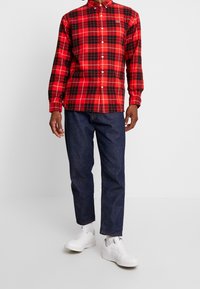 Camisa de cuadros rojo y negro, de botones, de manga larga, de material de algodón; combinada con pantalones de mezclilla oscuros y zapatillas blancas.