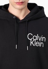 Black hoodie made of cotton, featuring a white "Calvin Klein" logo on the chest, adjustable drawstring with silver tips, and a kangaroo pocket.