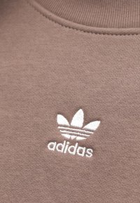 Καφέ φούτερ με ριμπ λαιμόκοψη που φέρει λευκό κεντημένο λογότυπο Adidas και τρεις ρίγες πάνω από το κείμενο. Μαλακή βαμβακερή υφή.