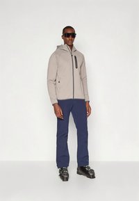 Man in een beige zip-up jack, marineblauwe broek, zwarte skibotten en grote zwarte zonnebril tegen een witte achtergrond.