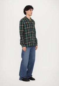 Grønn og svart rutete flanellskjorte med gule detaljer, knappeoppstramming, lange ermer; sammen med løse blå denimjeans og svarte sko.