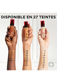 Des flacons de fond de teint avec des bouchons rouges tenus par trois mains ; des échantillons de différentes teintes de peau affichés sur des bras. Disponibles en 27 teintes.