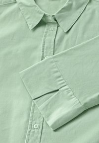 Ljusgrön skjorta i kordtyg med knappar, som har en spetsig krage, en enda knappknäppning och långa ärmar med manschetter och en slät yta.