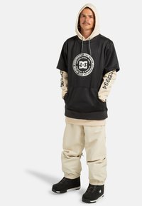 Felpa oversize nera e beige con logo, pantaloni beige e stivali neri. Tessuto morbido, cappuccio con cordini, tasche frontali e polsini elasticizzati.